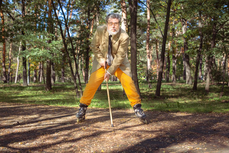 Funny old man is roller skating in the park. An active grandfather with a walking stick dances and makes faces. Grandfather with a white beard.の写真素材