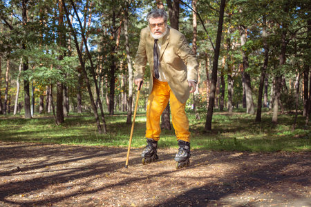 Funny old man is roller skating in the park. An active grandfather with a walking stick dances and makes faces. Grandfather with a white beard.の写真素材