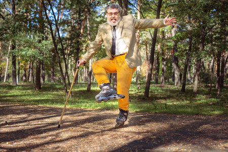 Funny old man is roller skating in the park. An active grandfather with a walking stick dances and makes faces. Grandfather with a white beard.の写真素材