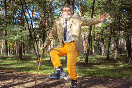 Funny old man is roller skating in the park. An active grandfather with a walking stick dances and makes faces. Grandfather with a white beard.の写真素材