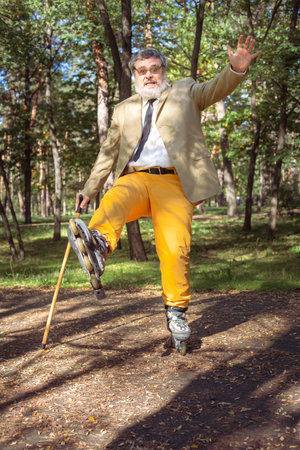 Funny old man is roller skating in the park. An active grandfather with a walking stick dances and makes faces. Grandfather with a white beard.の写真素材