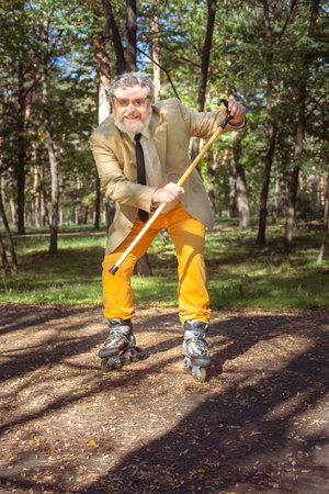 Funny old man is roller skating in the park. An active grandfather with a walking stick dances and makes faces. Grandfather with a white beard.の写真素材