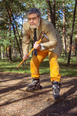 Funny old man is roller skating in the park. An active grandfather with a walking stick dances and makes faces. Grandfather with a white beard.の写真素材