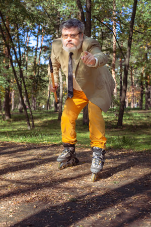 Funny old man is roller skating in the park. An active grandfather with a walking stick dances and makes faces. Grandfather with a white beard.の写真素材