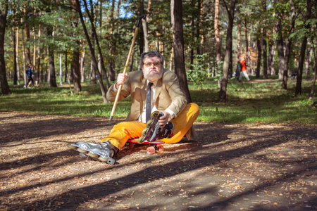 Funny old man is roller skating in the park. An active grandfather with a walking stick dances and makes faces. Grandfather with a white beard.の写真素材