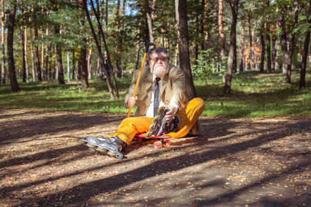 Funny old man is roller skating in the park. An active grandfather with a walking stick dances and makes faces. Grandfather with a white beard.の写真素材