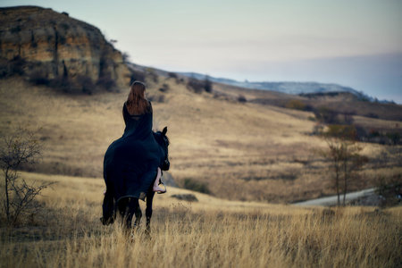 A young girl in a black cloak riding on her black horse, standing with her back turned in a field on...