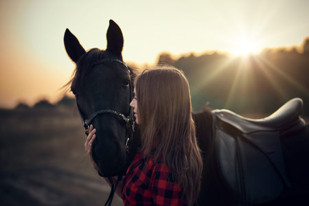 Young woman farmer in a red checkered shirt hugs her black horse at sunset - concept of love between...