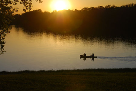 A canoe is paddled towards the setting sun over calm water.  They leave a small wake as their boat...