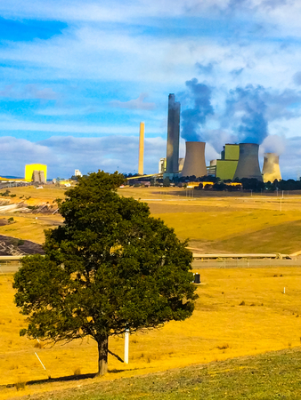 Coal powered electric energy generator plant in the middle of yellow desert emitting thick smoke to the clear air sunny blue skyの写真素材