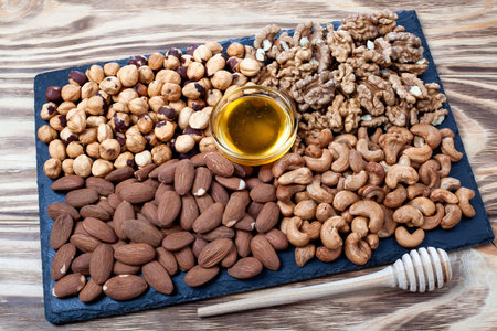 Various nuts sorted in glass bowl with honey bowl and honey dipper. Mixed nuts on wooden table. Black stone plate on wooden background.の写真素材