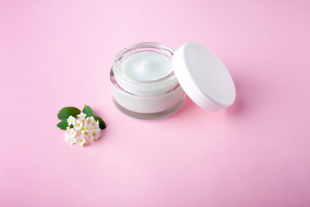 Glass jar and white cap with body cream on pink background with small white flowers. Transparent jar...