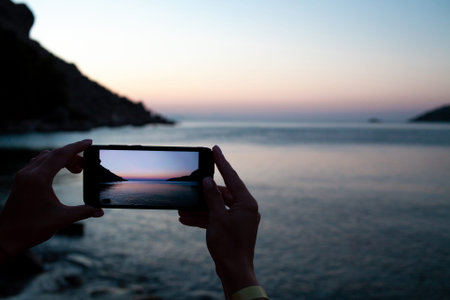 woman hands holding mobile phone at sunrise over sea bay. Taking photo of sunset over rocks in water...