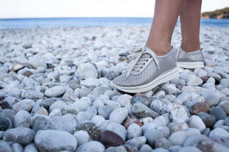 Silver leather sneakers shoe on a female leg walking on beach. Walking on pebble beach in comfortable shoes.の写真素材