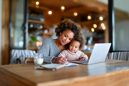 A mother helps her young child with learning tasks at a wooden table. They share a cozy moment with a laptop, notebook, and a glass of milk.の素材