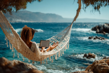 A woman relaxes in a hammock near the water, typing on her laptop while enjoying the sunny beach view and gentle waves.の素材