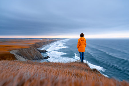 A person in an orange jacket stands on a cliff, gazing at the rolling ocean waves under a colorful sky during sunset. The scene captures nature's beauty.の素材