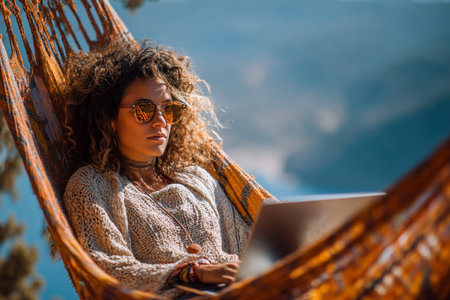 A woman enjoys her time in a cozy hammock, working on her laptop surrounded by stunning mountain views under a bright blue sky.の素材