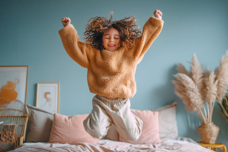 A child in a warm sweater leaps joyfully on a bed in a soothing room, surrounded by soft pillows and calming decor, creating a moment of pure happiness.の素材