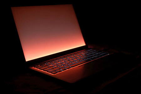 A laptop sits in a dark, minimal workspace with its glowing screen casting a warm light in the room. The atmosphere is calm and focused for work.の素材