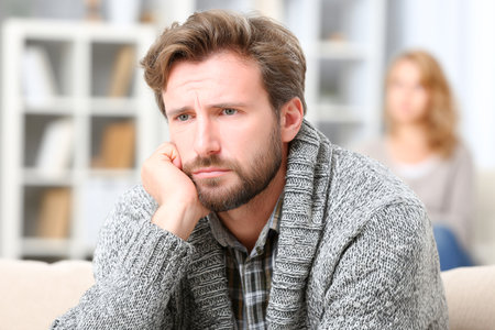 A man sits on a couch in a therapy room, showing nervous body language with a worried expression. In the background, a woman participates in the session.の素材