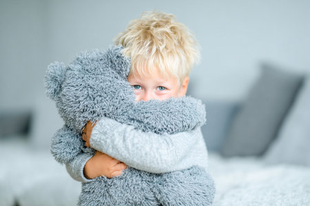 A young child with tousled hair hugs a soft gray stuffed animal tightly in a bright, cozy room filled with soft furnishings.の素材