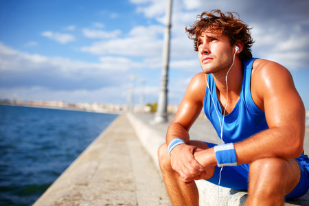 A runner sits by the water, enjoying a moment of relaxation with headphones on after an intense workout. The sky is clear and the sun shines brightly, creating a calm atmosphere.の素材