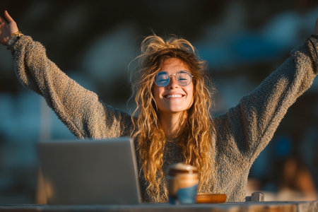 A happy freelancer celebrates a recent success while working outdoors. She raises her arms in joy, with a laptop and coffee cup beside her as the sun sets.の素材