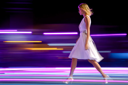 A model elegantly strides down a vibrant runway illuminated by colorful lights, showcasing a stylish dress in a lively fashion show atmosphere.の素材