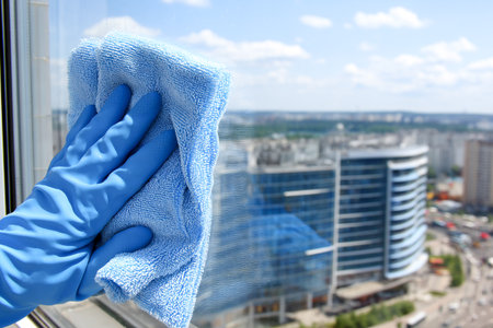 A person in blue gloves cleans windows using a cloth and spray. The bright sky and city skyline enhance the busy atmosphere of the scene.の素材