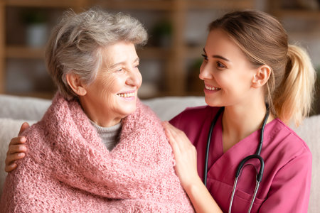 A care nurse smiles warmly while assisting a senior woman wrapped in a cozy blanket. The caring atmosphere showcases the bond between them in a comfortable home environment.の素材