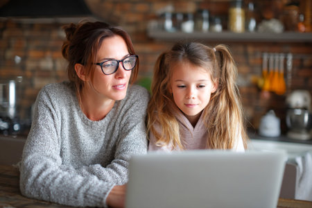 A female tutor guides a young girl through homework on a laptop in a warm and inviting home kitchen. The atmosphere is focused and nurturing.の素材