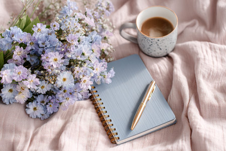 A light desk features a neatly placed notebook and pen alongside a coffee cup and fresh flowers. Soft fabric adds a cozy feel to the scene.の素材