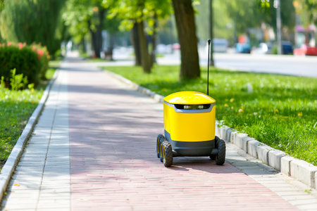 A delivery robot moves along a paved sidewalk, surrounded by trees and grass, as cars pass by in the background on a bright and clear day.の素材