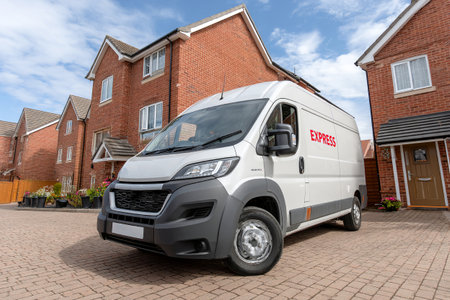 A delivery van with branding is parked in a quiet residential area. Homes with red brick facades surround the van under a bright blue sky.の素材