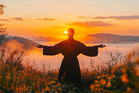 A person stands with arms open wide at sunrise, surrounded by mist-covered hills and tall grass. The warm colors fill the sky, creating a tranquil atmosphere.の素材