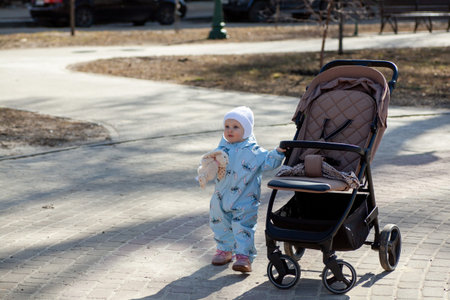 A little child pulls a stroller while holding a teddy bear. The setting is a park on a sunny day...