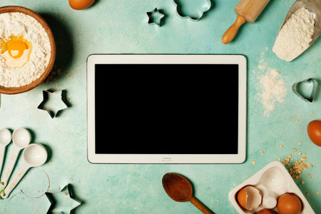 Baking background with flour, eggs, kitchen tools and tablet computer on blue rustic table. Top...
