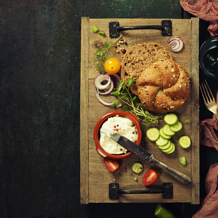 Homemade bread loaf and fresh ingredients for making vegetarian sandwiches (vegetables, herbs, cream cheese) on rustic dark background.の写真素材