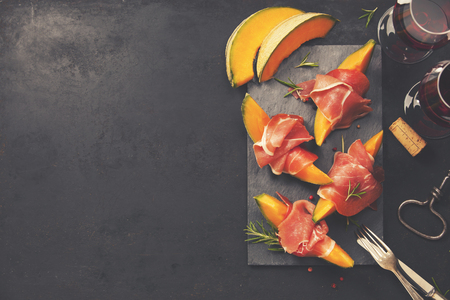 Prosciutto ham with cantaloupe melon, basil leaves and wine over dark grunge background. Top view, copy spaceの写真素材