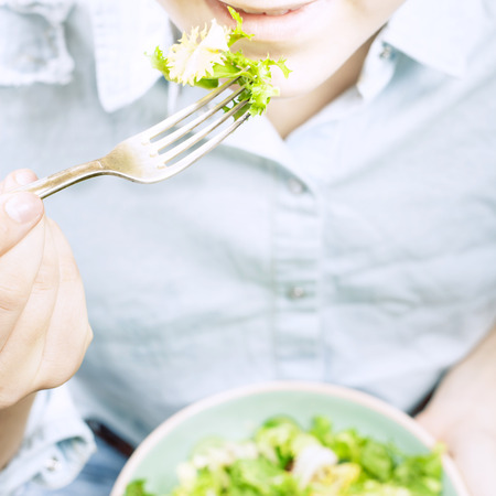 Fresh summer salad in bowl with lettuce, tomatoes, onions and cucumbers. Girl in jeans and blue shirt holding fork, top view. Clean eating, dieting food conceptの写真素材