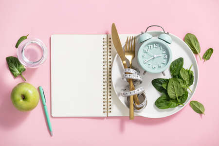 Composition with cutlery, measuring tape, blank paper notebook, spinach, apple, water and alarm clock on color background. Diet concept, copy spaceの写真素材