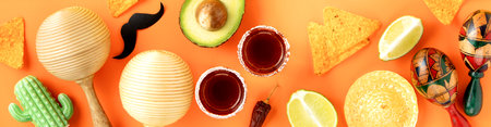 A vibrant Cinco de Mayo-inspired flat lay featuring a sombrero, maracas, tequila shots, lime, chili peppers, nacho chips on a bright yellow and orange backdropの写真素材