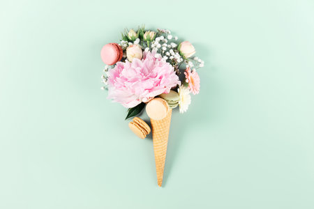Whimsical arrangement of fresh flowers and colorful macarons in a waffle cone with pastel sprinkles on mint green background.の写真素材