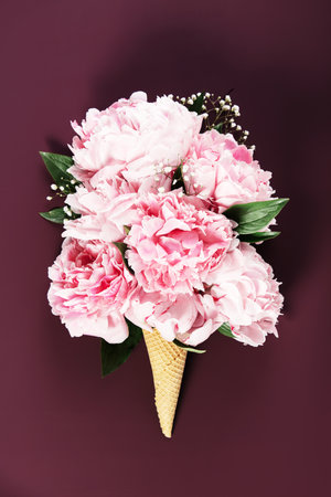 Artistic arrangement of pink peonies and greenery styled in a waffle cone on a rich burgundy backdrop.の写真素材