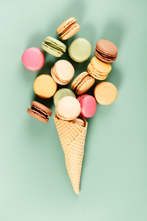 Creative flat lay of assorted macarons arranged in a waffle cone like ice cream, on a soft mint green background.の写真素材