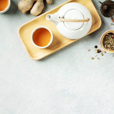 Top view of a white teapot and tea cup on a wooden tray with ginger root and loose-leaf herbal tea on a light stone background. Clean and natural wellness concept.の写真素材