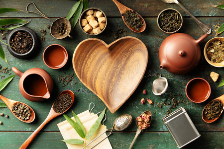 Heart-shaped wooden bowl surrounded by tea leaves, teaware, and natural ingredients on a textured green surface. Perfect for branding, wellness themes, and tea-related design projects.の写真素材