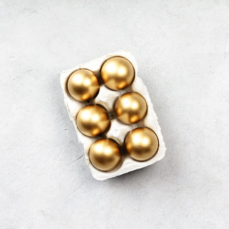 Six metallic golden eggs arranged in a recycled carton on neutral stone background. Minimal festive...