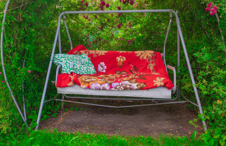 The horizontal photo shows garden swings, which are in the garden among flowers, grass, shrubs. On the swing is a blanket, a pillow. The veil on the red swing, the pillow is greenの写真素材